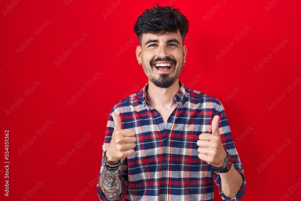 Young hispanic man with beard standing over red background success sign doing positive gesture with hand, thumbs up smiling and happy. cheerful expression and winner gesture. Young hispanic man with beard standing over red background success sign doing positive gesture with hand, thumbs up smiling and happy. cheerful expression and winner gesture.