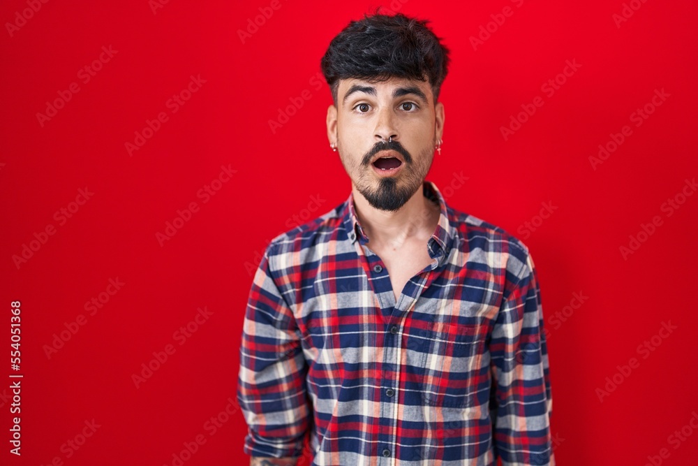 Young hispanic man with beard standing over red background afraid and shocked with surprise and amazed expression, fear and excited face. Young hispanic man with beard standing over red background afraid and shocked with surprise and amazed expression, fear and excited face.