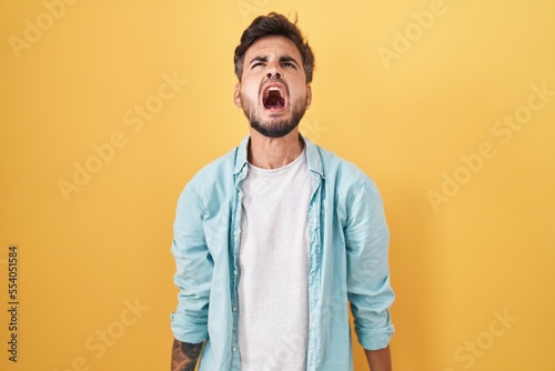 Young hispanic man with tattoos standing over yellow background angry and mad screaming frustrated and furious, shouting with anger. rage and aggressive concept. © Krakenimages.com
