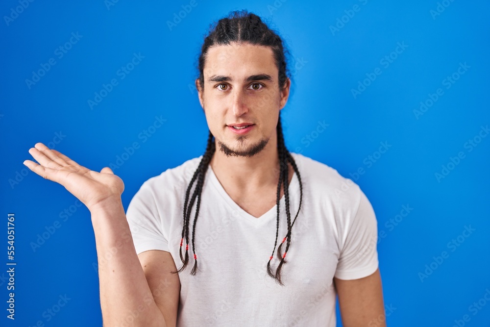 Hispanic man with long hair standing over blue background smiling cheerful presenting and pointing with palm of hand looking at the camera. Hispanic man with long hair standing over blue background smiling cheerful presenting and pointing with palm of hand looking at the camera.