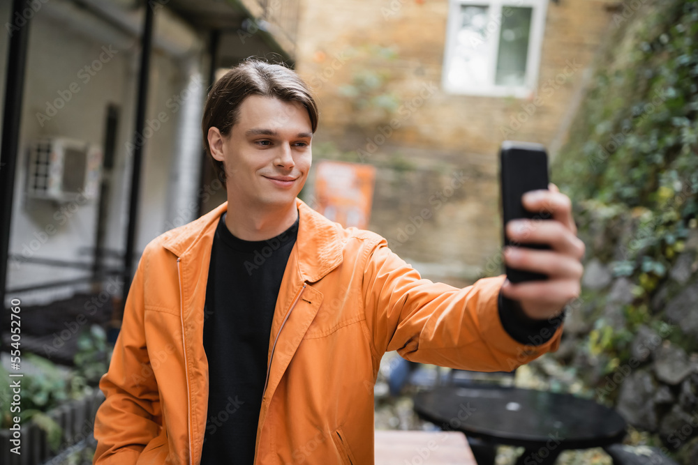 Smiling man in jacket taking selfie on smartphone on terrace of cafe. Smiling man in jacket taking selfie on smartphone on terrace of cafe.