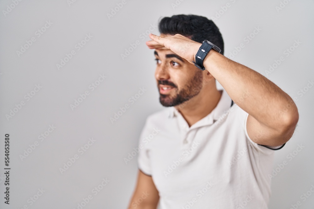 Young hispanic man with beard wearing casual clothes over white background very happy and smiling looking far away with hand over head. searching concept. Young hispanic man with beard wearing casual clothes over white background very happy and smiling looking far away with hand over head. searching concept.