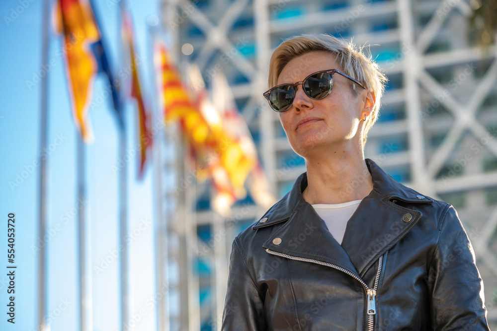 30s blond woman with short hair in sunglasses and leather jacket next to modern building in Barcelona with european flags on background