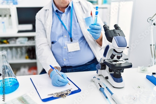 Middle age grey-haired man wearing scientist uniform holding test tube and write on document at laboratory photo