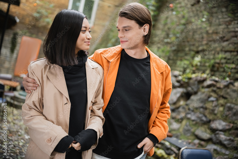 Young man hugging smiling asian girlfriend in trench coat in outdoor cafe. Young man hugging smiling asian girlfriend in trench coat in outdoor cafe.