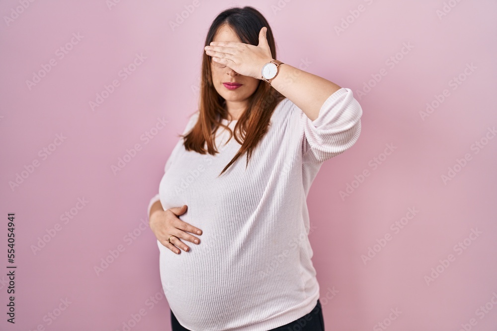 Fototapeta premium Pregnant woman standing over pink background covering eyes with hand, looking serious and sad. sightless, hiding and rejection concept
