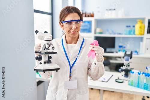 Young brunette woman working at scientist laboratory holding pink ribbon skeptic and nervous, frowning upset because of problem. negative person.