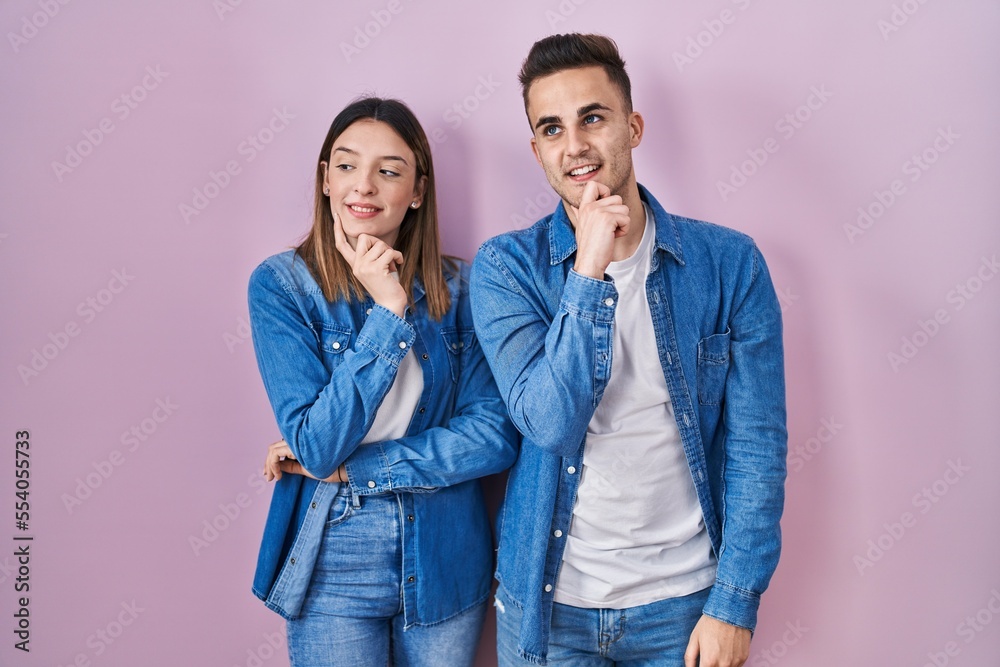 Young hispanic couple standing over pink background with hand on chin thinking about question, pensive expression. smiling and thoughtful face. doubt concept. Young hispanic couple standing over pink background with hand on chin thinking about question, pensive expression. smiling and thoughtful face. doubt concept.