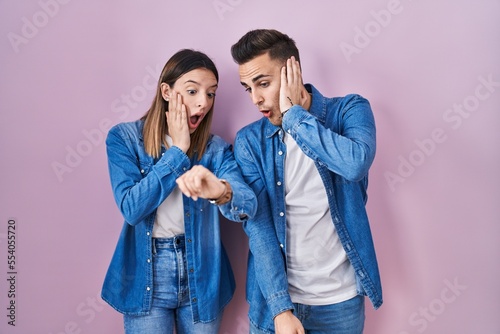 Young hispanic couple standing over pink background looking at the watch time worried, afraid of getting late