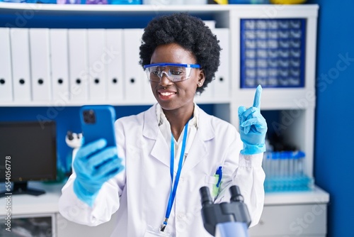 African young woman working at scientist laboratory doing video call smiling happy pointing with hand and finger to the side
