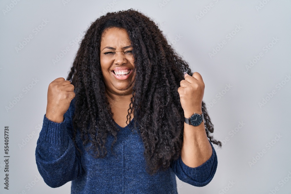 Plus size hispanic woman standing over white background very happy and excited doing winner gesture with arms raised, smiling and screaming for success. celebration concept. Plus size hispanic woman standing over white background very happy and excited doing winner gesture with arms raised, smiling and screaming for success. celebration concept.