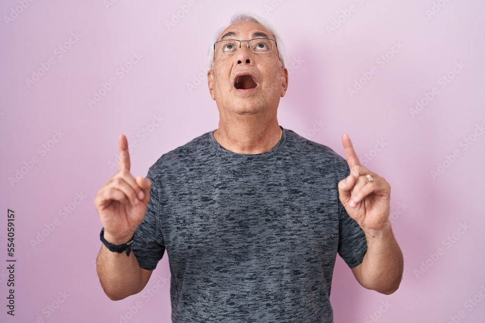 Middle age man with grey hair standing over pink background amazed and surprised looking up and pointing with fingers and raised arms. Middle age man with grey hair standing over pink background amazed and surprised looking up and pointing with fingers and raised arms.