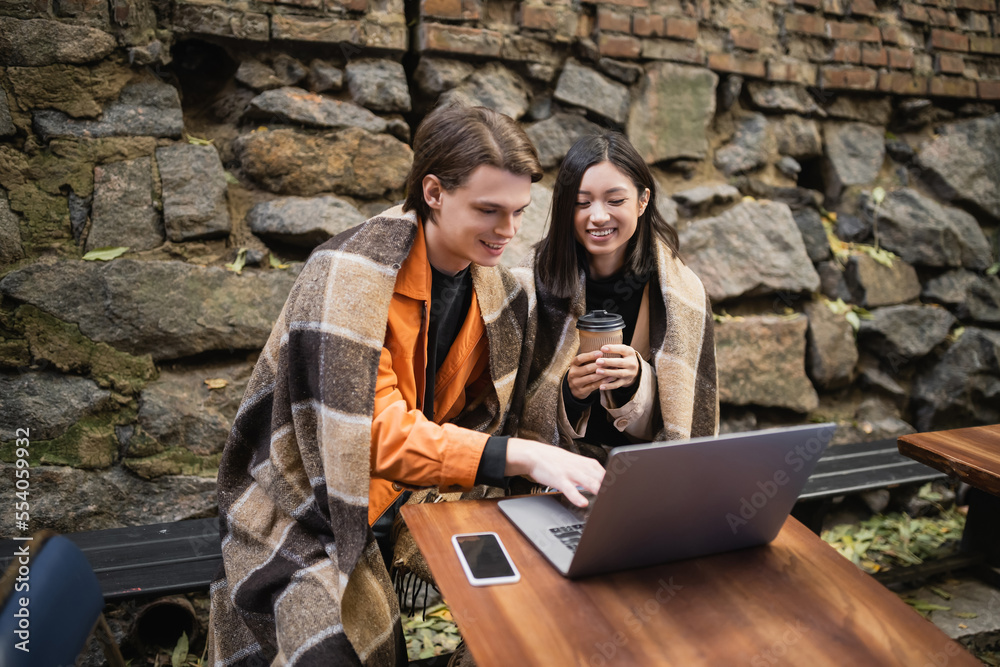 Positive multiethnic couple in blanket using laptop on terrace of cafe. Positive multiethnic couple in blanket using laptop on terrace of cafe.
