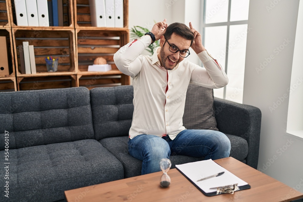 Young hispanic man with beard working at consultation office posing funny and crazy with fingers on head as bunny ears, smiling cheerful