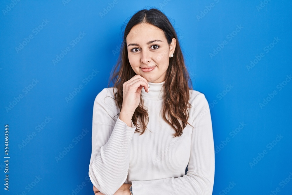 Young hispanic woman standing over blue background with hand on chin thinking about question, pensive expression. smiling and thoughtful face. doubt concept.