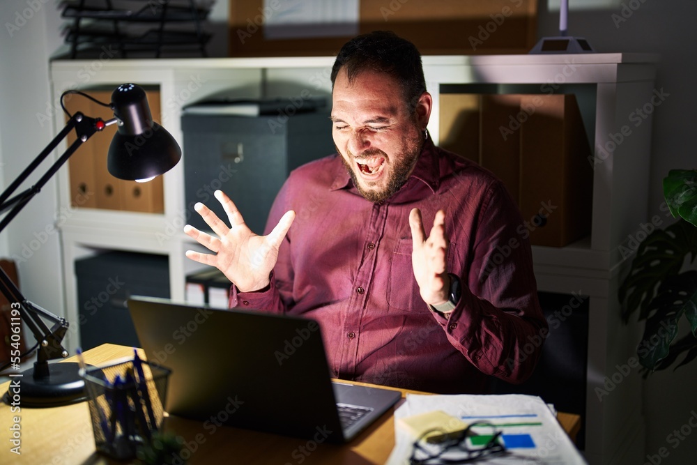 Plus size hispanic man with beard working at the office at night celebrating mad and crazy for success with arms raised and closed eyes screaming excited. winner concept Plus size hispanic man with beard working at the office at night celebrating mad and crazy for success with arms raised and closed eyes screaming excited. winner concept