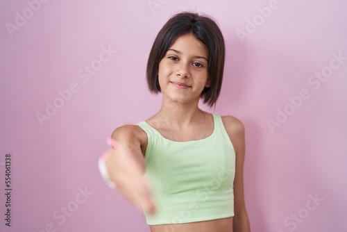 Young girl standing over pink background smiling friendly offering handshake as greeting and welcoming. successful business. © Krakenimages.com