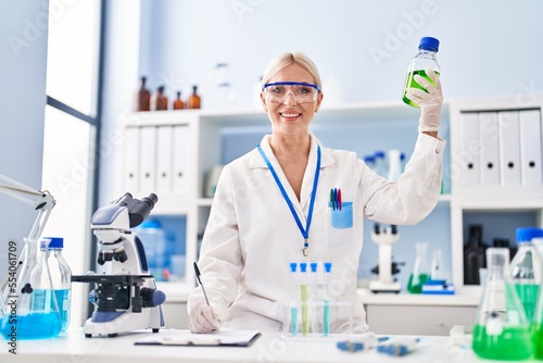 Young blonde woman wearing scientist uniform holding bottle at laboratory