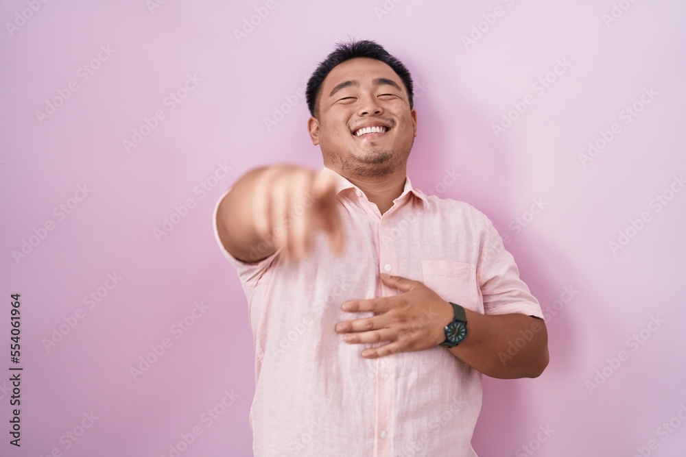 Chinese young man standing over pink background laughing at you, pointing finger to the camera with hand over body, shame expression Chinese young man standing over pink background laughing at you, pointing finger to the camera with hand over body, shame expression