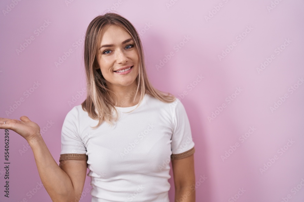 Fototapeta premium Blonde caucasian woman standing over pink background smiling cheerful presenting and pointing with palm of hand looking at the camera.