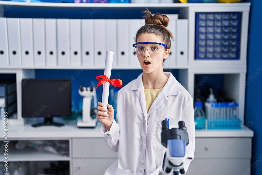 Teenager girl working at scientist laboratory holding degree scared and amazed with open mouth for surprise, disbelief face