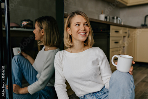 incredible smiling young woman with short blond hair wearing white shirt and jeans sitting at home with coffee and waiting for friends 