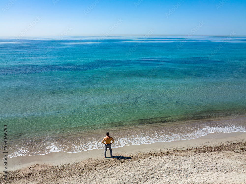 Veduta aerea di una spiaggia con un mare cristallino - si vede un uomo in solitaria che guarda il mare. Veduta aerea di una spiaggia con un mare cristallino - si vede un uomo in solitaria che guarda il mare.