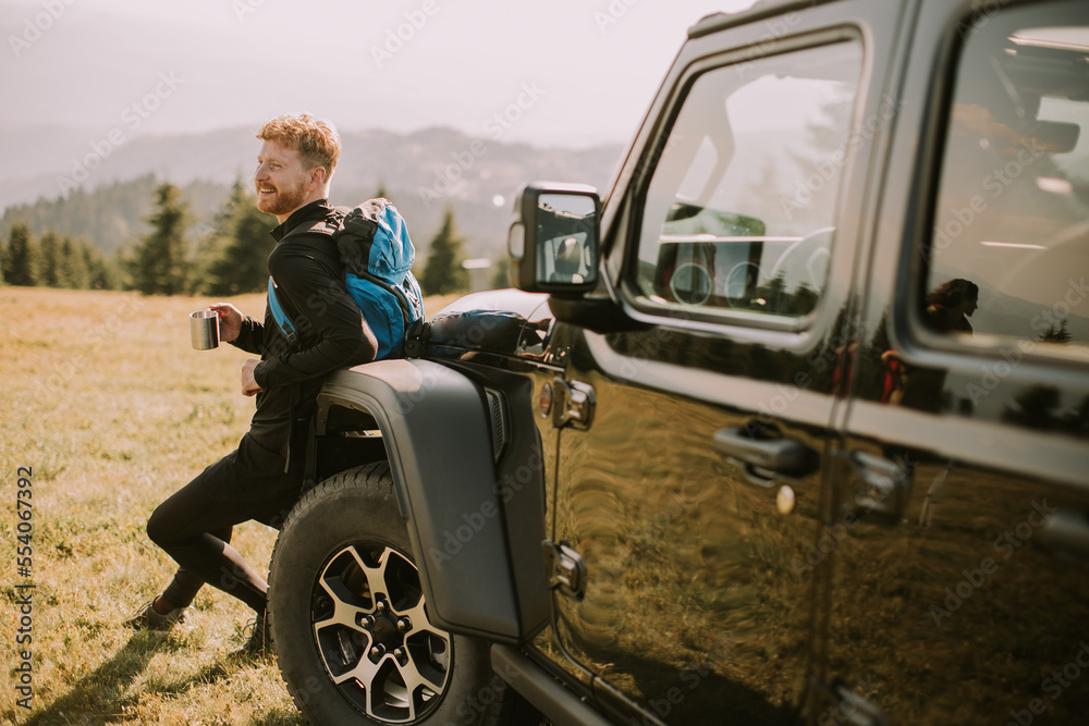 Young man relaxing and drinking coffee by the terrain vehicle hood at countryside