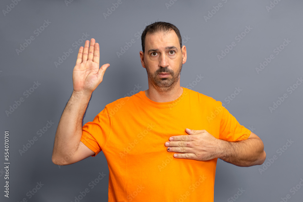 Bearded hispanic man wearing orange t-shirt isolated on gray background swearing, putting hand on chest Bearded hispanic man wearing orange t-shirt isolated on gray background swearing, putting hand on chest