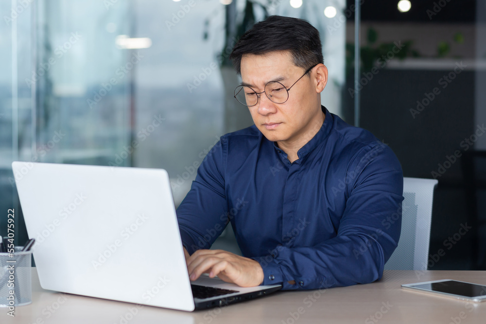 Serious thinking asian businessman working inside modern office, mature man in shirt and glasses using laptop at work, investor pondering complex decision. Serious thinking asian businessman working inside modern office, mature man in shirt and glasses using laptop at work, investor pondering complex decision.