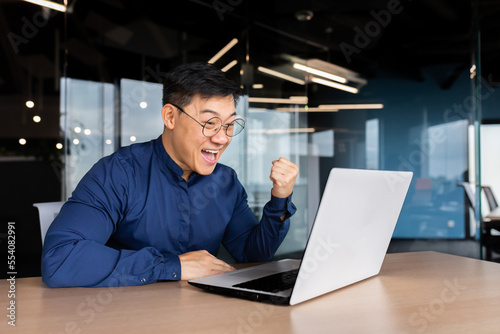 Successful asian man celebrating victory and success received news of good achievement online, businessman looking at laptop screen and holding hand up triumph gesture, worker inside office.