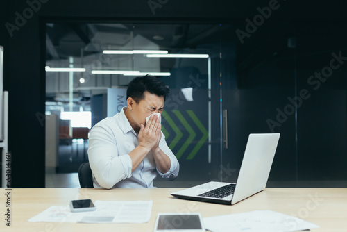 Illness at work. A young Asian man sits in the office at the table, wipes his nose with a napkin, has a runny nose, flu, feels bad, is exhausted.