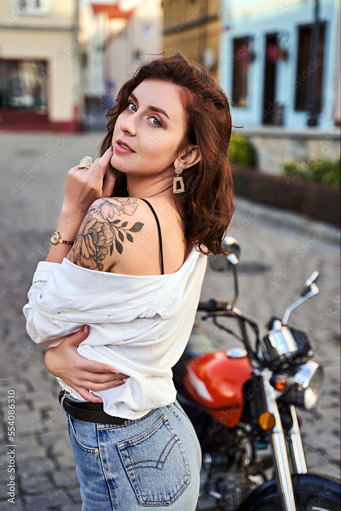 Young beautiful woman shows her bare shoulder with a tattoo standing by a bike on the street
