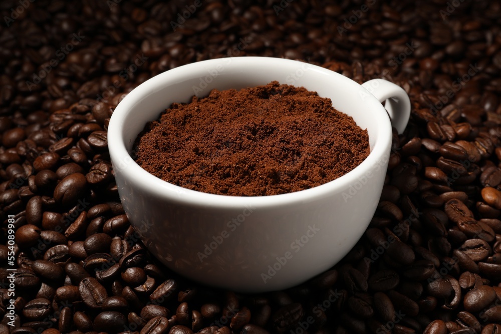 Cup with ground coffee on roasted beans, closeup Cup with ground coffee on roasted beans, closeup