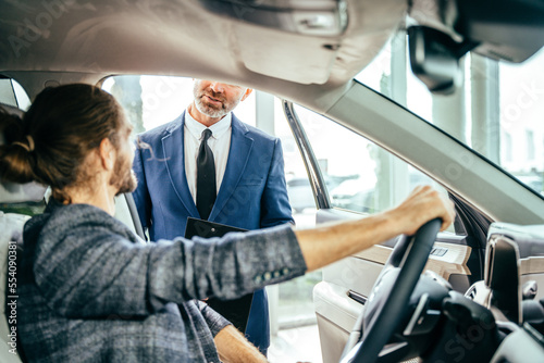 Serious dealer looking at pensive business man sitting in car to checking features. Young successful customer man deciding to buy new auto in showroom.