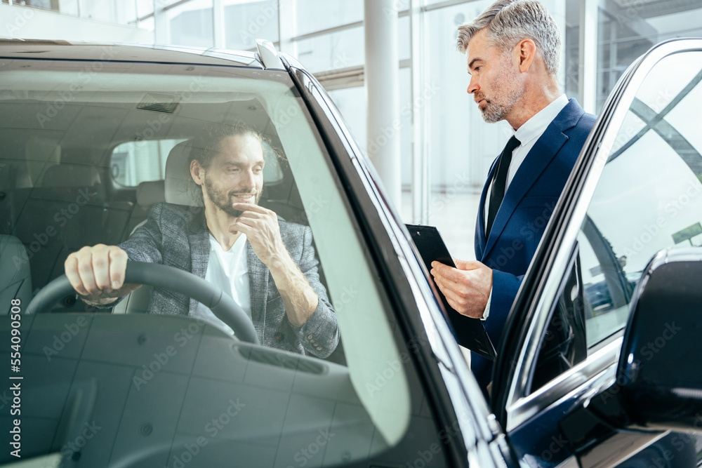 Seller stands at white car and holds tablet in hands. He point on it. Another man sits in car and holds insurance paper. He is serious and concentrated. They have conversation. Seller stands at white car and holds tablet in hands. He point on it. Another man sits in car and holds insurance paper. He is serious and concentrated. They have conversation.