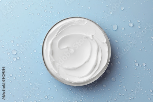 Jar of face cream on light blue surface covered with water drops, top view