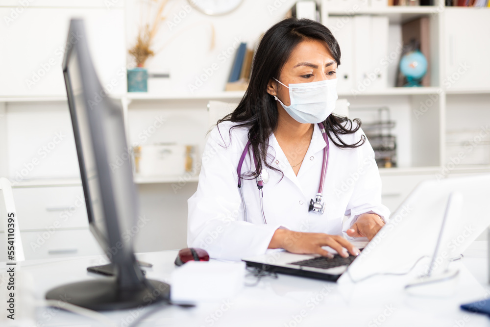 Experienced female physician in protective mask filling up medical forms on laptop while sitting at table in office - obrazy, fototapety, plakaty 