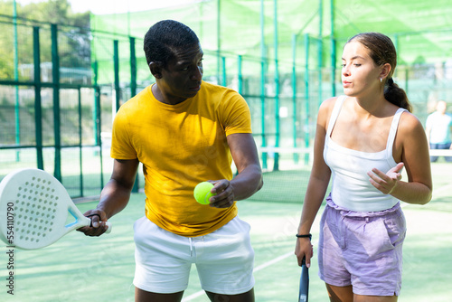 African-american man padel trainer teaching young caucasian woman to serve ball on outdoor court.