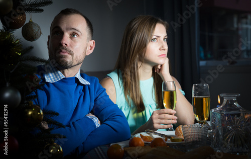 Bored or quarreled young man and woman sitting at holiday table and watching tv at Christmas night