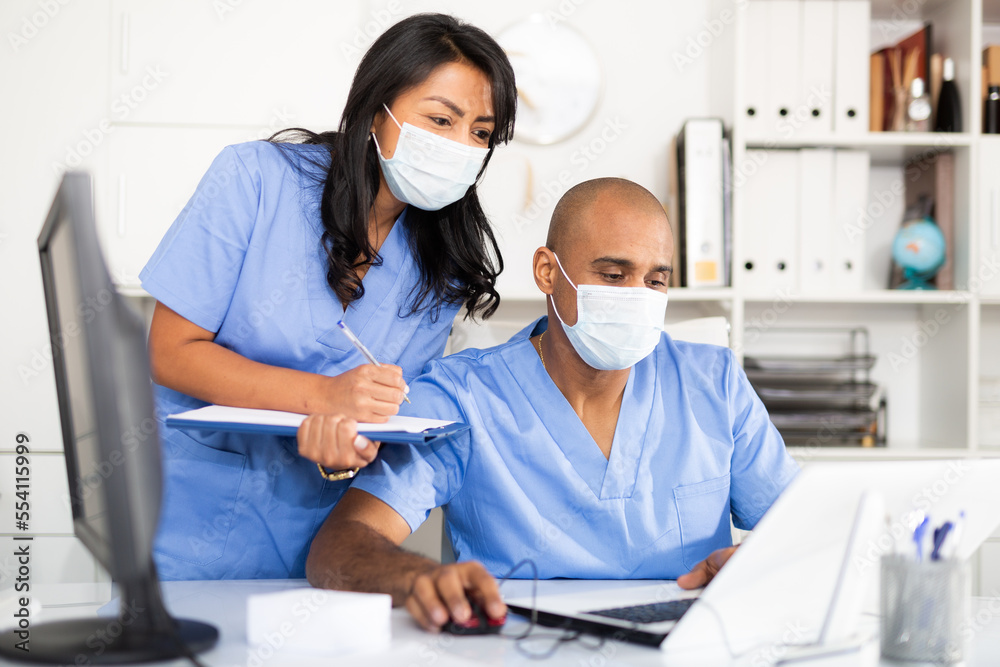 Two focused physicians wearing medical masks working with case histories on laptop in modern office