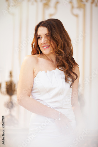 Beautiful charming senior woman in festive luxury white dress on the vintage background with antique table in gold tones. A brunette  model with long hair and bright smile 