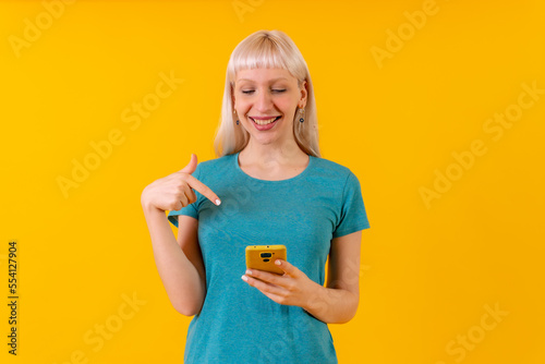 Smiling pointing at the phone, blonde caucasian girl in studio on yellow background