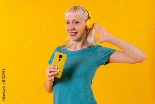 listening to music with headphones online, blonde caucasian girl in studio on yellow background