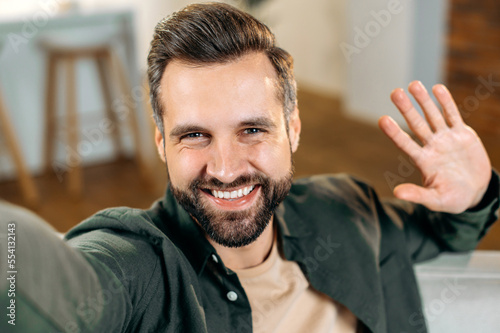 Cheerful attractive positive caucasian bearded man, in a casual shirt, takes a selfie on his smartphone, waves hand, looks into the front camera of the mobile phone, smiles friendly