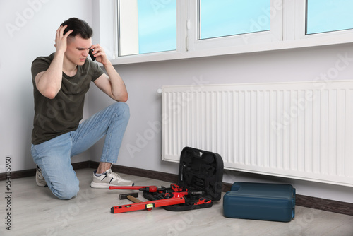 Handyman with pipe wrench talking on phone near radiator in room