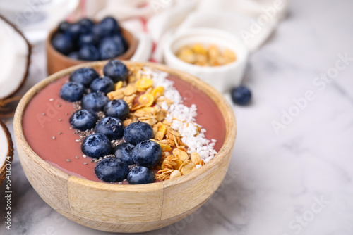 Bowl of delicious fruit smoothie with fresh blueberries, granola and coconut flakes on white marble table, closeup. Space for text