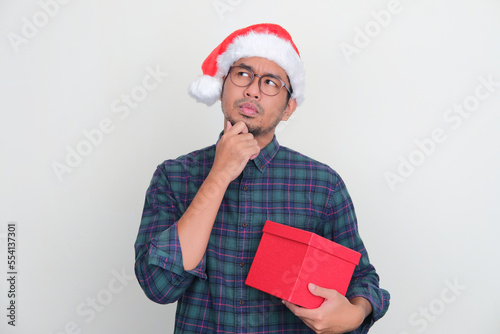 Adult Asian man wearing Christmas hat and holding gify box showing thinking expression photo