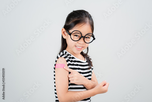 Asian little girl wear glasses showing her arm with bandage after virus vaccine happy Kid and child vaccination campaign. Protect your kid with vaccine concept