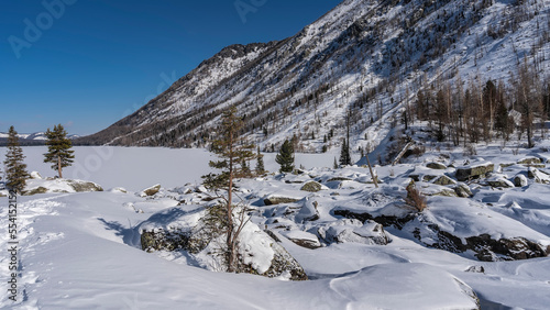 Picturesque boulders under a layer of snow on the shore of a frozen lake. A chain of footprints is trampled in snowdrifts. Mountains against the blue sky. Altai. Lower Multinskoe Lake